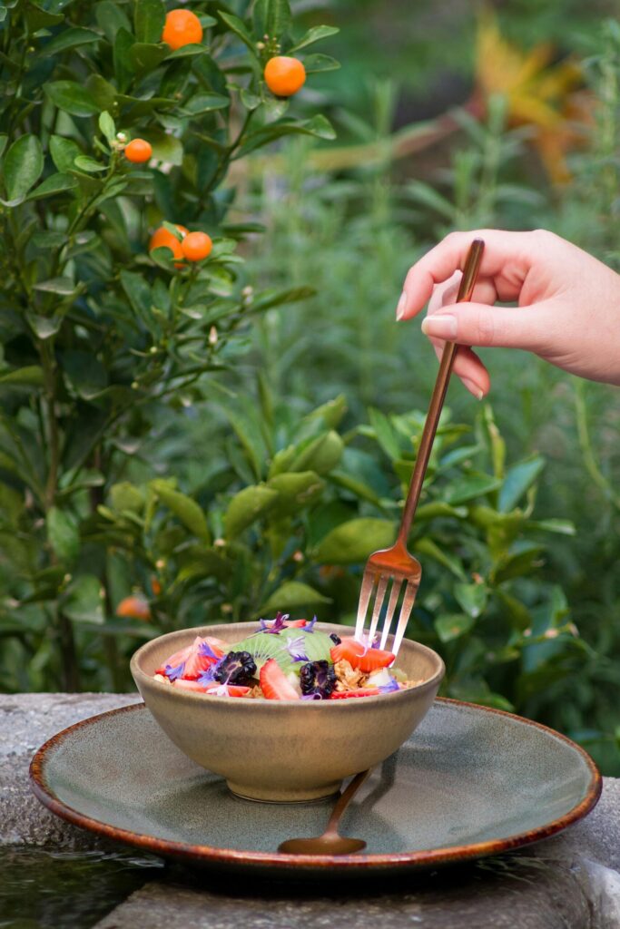 A hand holds a fork over a colorful salad, set outdoors by a citrus tree.