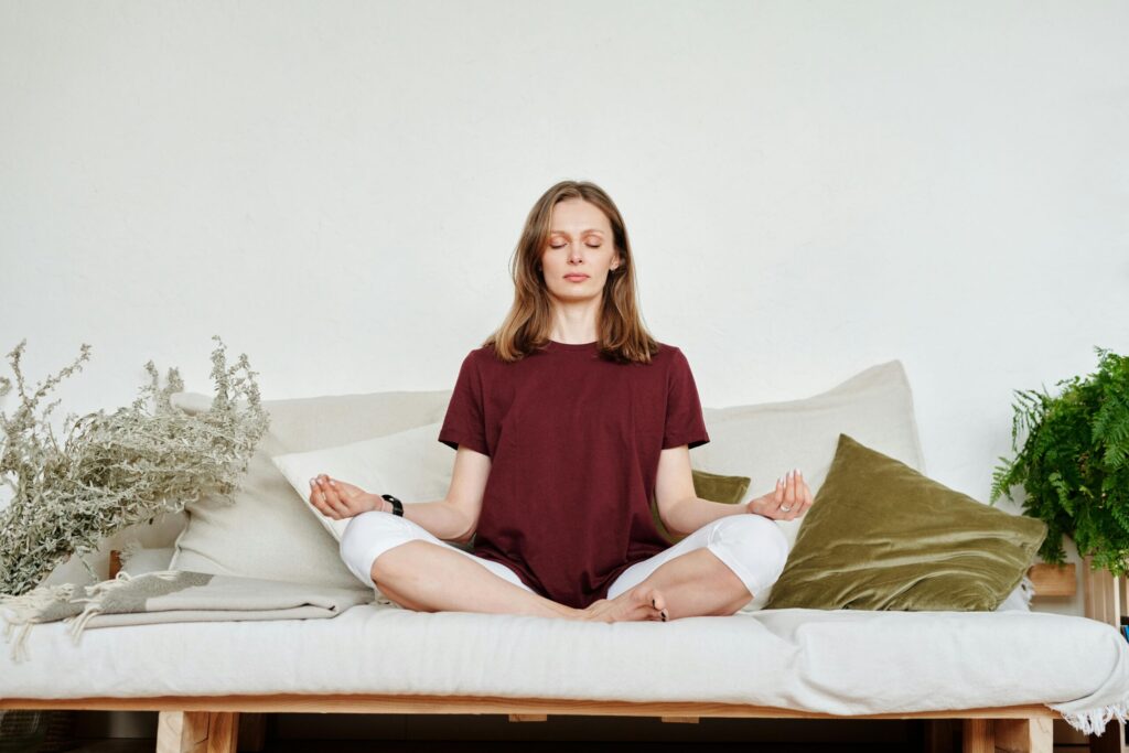 Woman meditating indoors on a sofa, exuding calmness and mindfulness, surrounded by greenery.