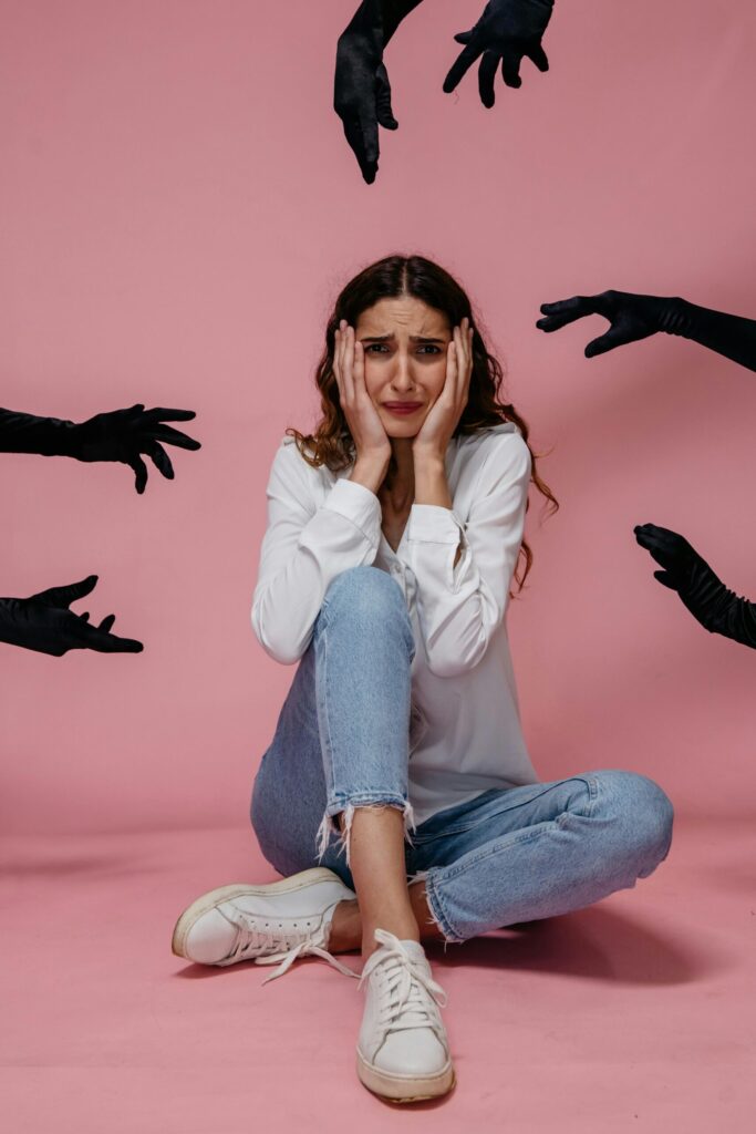 A woman looks anxious and stressed, sitting against a pink backdrop with hands reaching towards her.